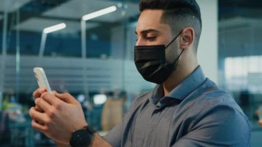 Multiracial Indian ethnic man guy businessman entrepreneur worker manager employer executive in medical face protective mask in office chatting texting in mobile phone browsing online smartphone app