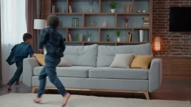 Happy two energetic little preschool kids siblings brothers African American ethnic children friends having fun running around couch sofa at home living room playful active together run game playing