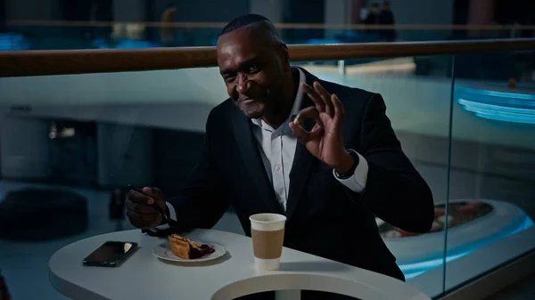 Ethnic African American middle-aged businessman entrepreneur man visitor sitting at cafe table eating pie good tasty cake dessert showing thumb up ok okay gesture recommend satisfied in restaurant