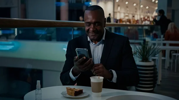 Mature adult middle-aged ethnic African American businessman man sitting in cafe with coffee tea and slice of pie cake make photo selfie with mobile phone camera posing for smartphone image shooting