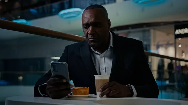 African American adult middle-aged businessman 50s ethnic man male employer entrepreneur sitting at table in cafe drink coffee drinking tea browsing mobile phone smartphone app talk tell video call