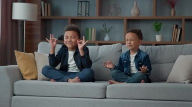 Two funny little African American ethnic children boys brothers siblings kids meditating together sitting on couch in lotus position. Small schoolboys meditate. Spiritual development yoga meditation