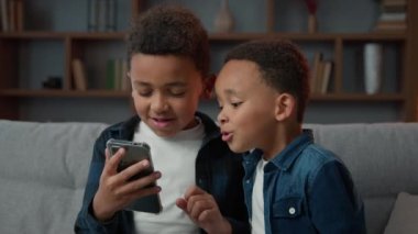 Two funny little ethnic African American children kids siblings brothers boys friends with phone smartphone playing mobile game browsing social media at home having fun with cellphone app sit on sofa