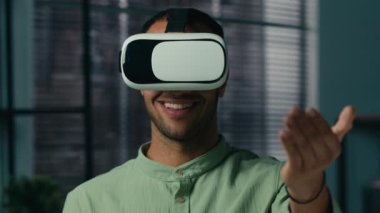 Close-up smiling latin american man wearing virtual reality glasses friendly showing invitation gesture asking to come welcome symbol making choice male player playing in cyberspace with vr goggles