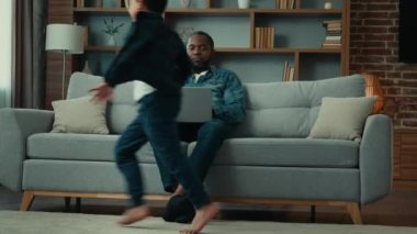 Stressed African American ethnic father man freelancer businessman at home sitting on couch work with laptop talking phone noisy active children sons little boys kids running around sofa run playing
