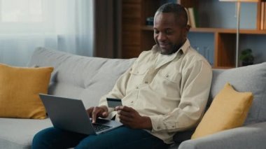 African american middle aged man sitting on couch mature male customer paying purchase credit card on laptop makes order online store pays using banking computer application successful remote payment