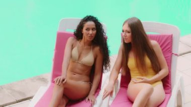 Smiling tanned multiracial girls women friends in swimsuits rest on sun loungers talk gossiping cheerful millennial hispanic and caucasian female students tourists girlfriends enjoy summer fun by pool