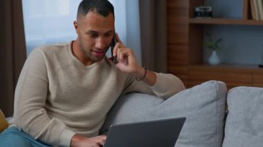 African businessman work freelance at home sit on couch american latino adult multiracial man talking mobile phone chatting online on computer multitasking communicate with client distant make order