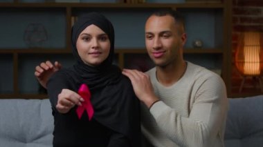 Multiracial family muslim woman and african american man sit on sofa indoors hold red ribbon prevent symbol HIV AIDS protect immune deficiency syndrome blood donation health care and world cancer day