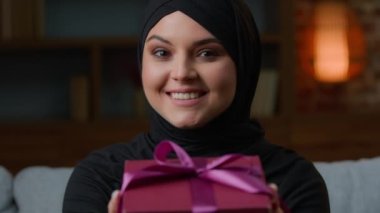 Smiling woman in hijab hold pink gift box with ribbon wishing happy birthday Christmas. Islamic muslim saudi girl young female give holiday present congratulating with anniversary festive celebration
