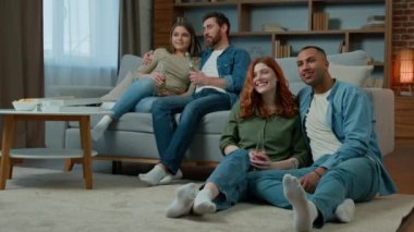Multiracial friends men women watching television at home hispanic couple relax on couch african american caucasian family sit on floor enjoy TV comedy film. Diverse ethnic people laughing drink beer