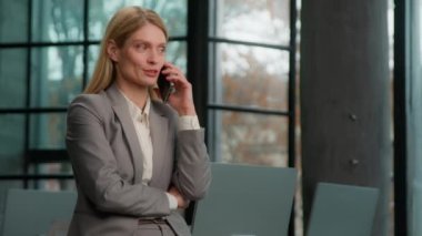 Caucasian middle-aged adult lady in office chat with colleague using mobile phone listen to news answer call. Woman executive businesswoman CEO female talking speak on telephone indoors business talk