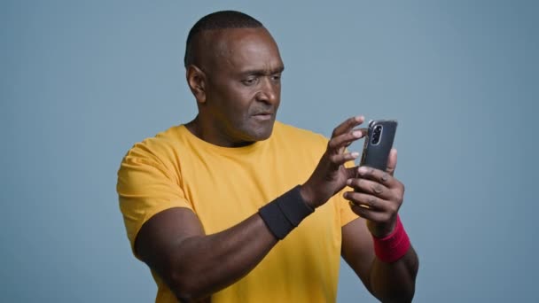 成熟的非洲运动员站在演播室灰色背景下看手机屏幕上的男性运动员教练浏览运动申请 展示认可的手势 — 图库视频影像