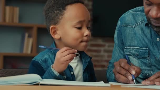 アフリカ系アメリカ人の子供で 父親は家庭で学ぶ パパの先生助ける小さな息子の生徒準備のためのテスト小学校の子供を行う宿題とともにパパ男書きタスク遠隔教育 — ストック動画