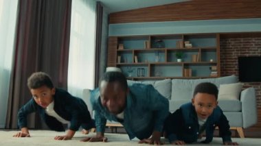 African American strong father man dad with two cute little sons teaching children siblings kids push-ups at floor in home living room teach boys press-up sport exercise fitness workout game together