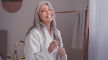 Old Caucasian smiling laughing lady 60s female smile laugh happy senior mature woman looking at camera holding hands to side having fun in bathroom pampering skin wrinkle care spa anti age hygiene