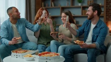 Happy diverse friends sitting on couch clinking bottles of beer celebrating birthday event friday home party. Excited multiracial african hispanic people men women eating pizza drinking alcohol dinner