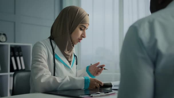 イスラム教徒のアラビア人女性若い女性医師セラピストは 病気の患者のための医療処方レシピを書きますチェックリストに記入注意事項病気の症状は 診療所で男性のクライアントに健康保険を示しています — ストック動画