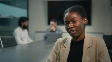 Webcam view African American ethnic woman business coach businesswoman talking video call conference chat looking at camera sitting at table on office on blurred background of talking men colleagues