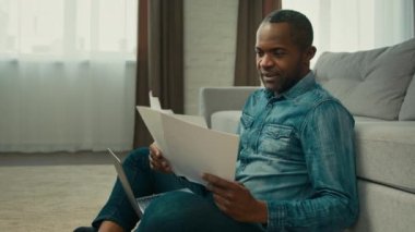 Mature african american man sitting in living room reading documents ethnic businessman freelancer working with papers analyzes finance checking report entering data in laptop work corporate paperwork