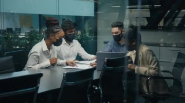 Diverse professional multiethnic business team workers colleagues in medical masks in office boardroom working with project papers discuss ideas results analyzing paperwork view from behind glass wall