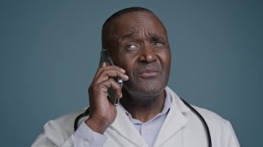 Close-up mature african american man talking mobile phone in studio gray background confused puzzled doubting male doctor in medical gown communicates with patient using smartphone hesitant answers