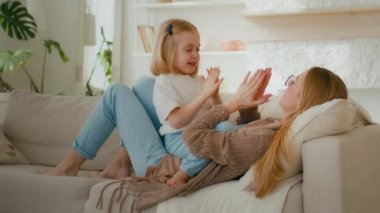 Happy parent Caucasian mom mother with cute kid daughter child girl baby playing patty cake game at home clapping hands lying on sofa couch at home living room carefree family having fun play together