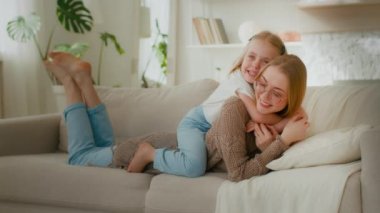 Caucasian mother in glasses lying on couch sofa in home living room little cute daughter kid child baby girl embrace hug cuddle mom by neck piggyback laughing together relaxing enjoy hugging cuddling
