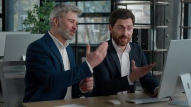 Two businessmen sitting in office looking at computer screen discussing growth sales on Internet rejoicing at lucrative contract discussing development trading happy men congratulations shake hands