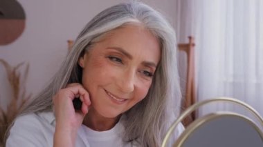 Anti-aging anti-wrinkled aging cosmetology. Old Caucasian woman senior mature aged lady female with perfect smooth complexion facial soft skin beautiful gray hair looking at mirror reflection smiling