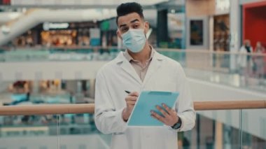 Ethnic Indian inspector scientist doctor medical male worker man writing in papers documents expertise check health safety rules supervisor with medical mask robe inspecting shopping mall write data