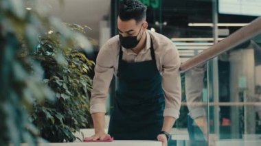 Hispanic Ethnic Indian man worker male waiter in medical mask catering worker in apron wipes table busy with cleaning before opening restaurant cafeteria pub prepares cafe for open finish work day