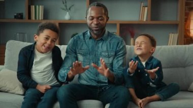 African American father man dad with two little children kids boys sons siblings with closed eyes sitting at couch meditate together doing breathing exercise breath calm meditation family meditating