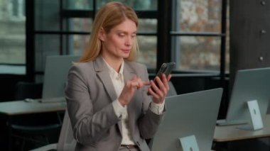 Caucasian middle-aged adult woman standing in office with cellphone browsing scrolling smartphone. 40s businesswoman using internet social media. Female employee take break use mobile phone online app