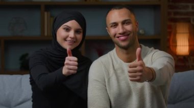 Smiling couple family african american man and arabian muslim woman diverse wife husband sitting on sofa together showing thumbs up like positive recommendation good attitude approve symbol of consent