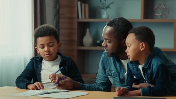 アフリカ系アメリカ人の父と2人の小さな息子の男の子子供がテーブルに座ってお父さんが教えるのを助ける生徒の男の子の息子は宿題の勉強を手伝ってレッスンクラスを書くホームスクールを学ぶ — ストック動画