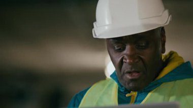 Close-up mature african american man engineer developer technician builder in protective helmet workwear using laptop indoors develop construction business project ponder work plan inspect speak idea