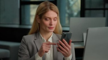 Caucasian middle-aged adult woman office worker executive businesswoman entrepreneur indoors scrolling browsing mobile phone shock amazed pointing at smartphone showing thumb up recommend good news