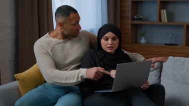 Married couple sit on sofa use laptop device choosing new apartment on internet website multiracial family shocked amazed diverse man woman search information by computer read good news talking indoor