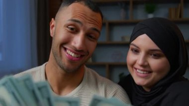 Rich multiracial couple spouses African American man counting money dollars with Arabian muslim woman in hijab planning family budget diverse partners considers finances cash banknotes sitting on sofa