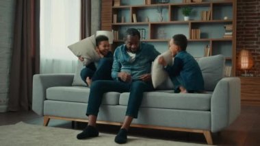 African American ethnic man father dad sitting at couch at home little noise active boys children sons play game on sofa hyperactive kids fighting pillows having fun fight difficulties of upbringing