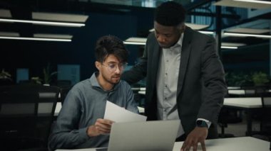 African american boss businessman mentor teaching indian man trainee discussing business project on papers explaining help with documents. Multiracial diverse ethnic men colleagues teamwork in office