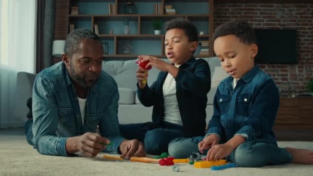 2人の小さな就学前の息子の男の子とアフリカ系アメリカ人の男性の父親子供たちはおもちゃで遊んでいる子供たちと家のお父さんで床に横たわってゲームをプレイ一緒に楽しい週末を楽しむ — ストック動画