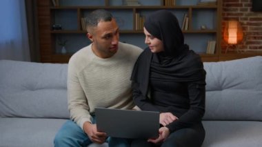 30s couple use laptop sitting on cozy sofa multiracial boyfriend african american man with muslim woman girlfriend shopping on internet watching funny video shopping online resting together at home