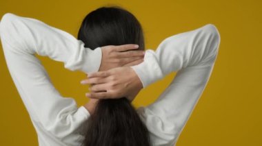Back view woman with wavy shiny curls hairdo long healthy beautiful hair stylish hairstyle makes ponytail out of her head female Indian ethnic girl model turning to camera in studio yellow background