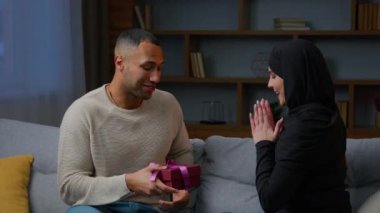 African american man boyfriend give surprise present pink birthday gift to loving girlfriend muslim woman in hijab romantic date multiracial couple family friends celebrate Valentine love day at home