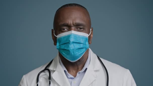 深刻なアフリカ系アメリカ人の成熟した男性医師の外科医医療ガウンと保護マスクでセラピストスタジオでポーズカメラを見て灰色の背景医療サービス医療保護コンセプト — ストック動画