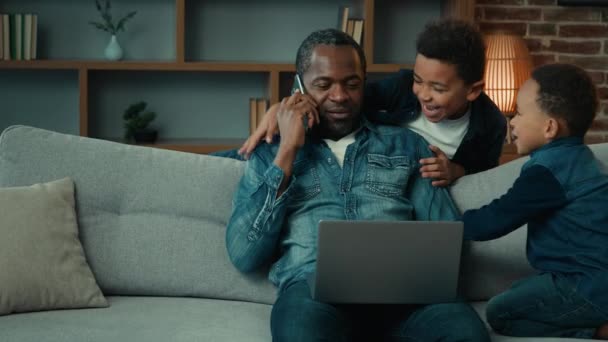 アフリカ系アメリカ人アメリカ人の父男フリーランスビジネスマンに自宅で座っていますソファワークとともにラップトップ電話騒々しい子供息子小さな男の子子供感動お父さんの需要注意気をそらすから仕事 — ストック動画