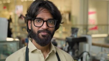 Successful small business Indian Arabian ethnic owner bearded man wearing eyeglasses standing in workplace cafeteria restaurant. Portrait of multiracial male cafe waiter worker guy looking at camera