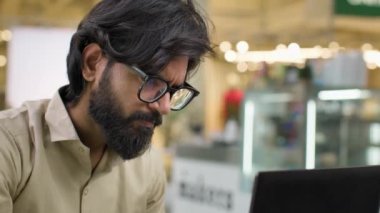 Adult concentrated Indian Arabian ethnic multiracial businessman trader man entrepreneur male manager in eyeglasses working with laptop computer thinking analyzing online trading finance data in cafe